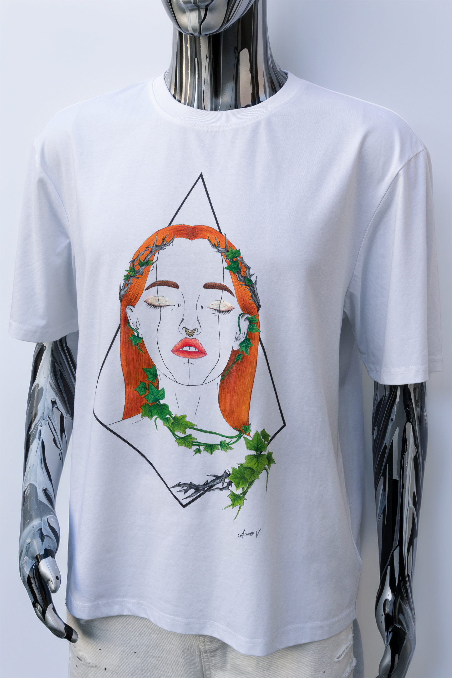 Mannequin wearing a white t-shirt with a graphic design on a plain background. The design is hand painted and shows a girl with orange hair, ivy leaves, and shimmery makeup.