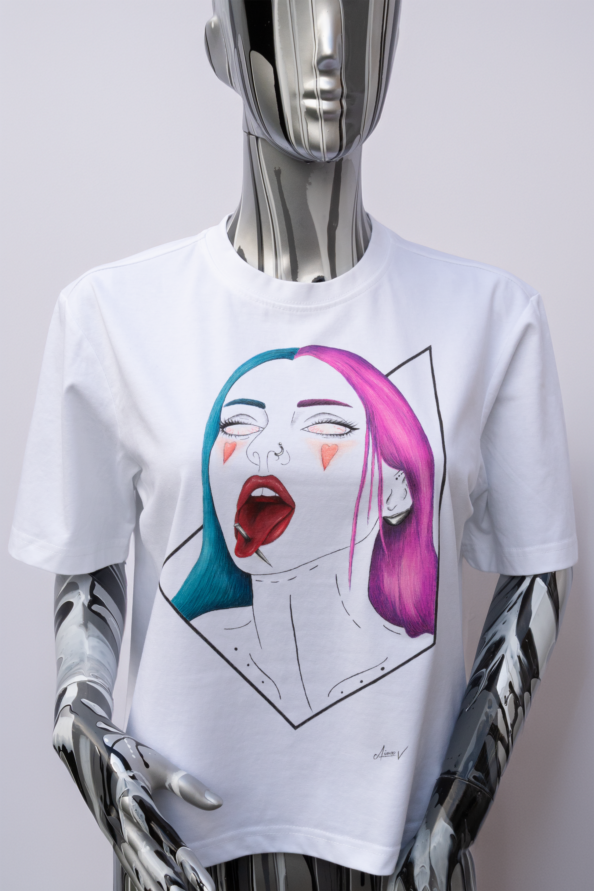 Mannequin wearing a white t-shirt with a graphic design on a plain background. The design is hand painted and shows a girl with half hair pink, half blue, 2 red hearts on the cheeks, and a needle through her tongue.
