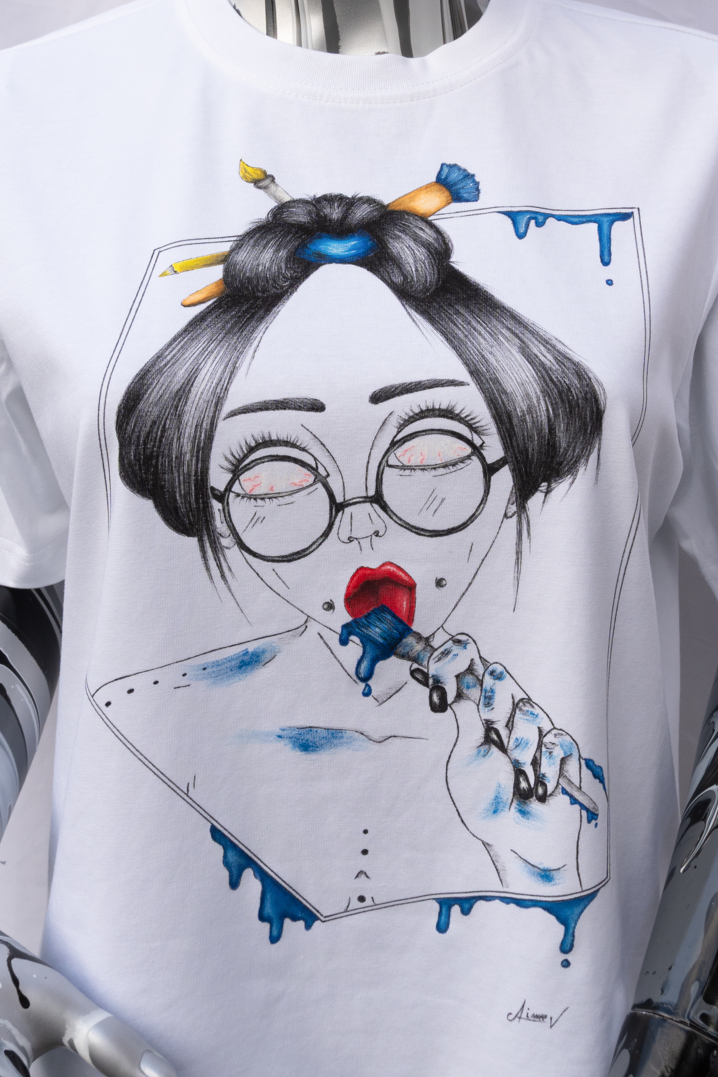 A close view of a mannequin wearing a white t-shirt with a graphic design on a plain background. The design is hand painted and shows a girl with round glasses, blue paint dripping and a brush.