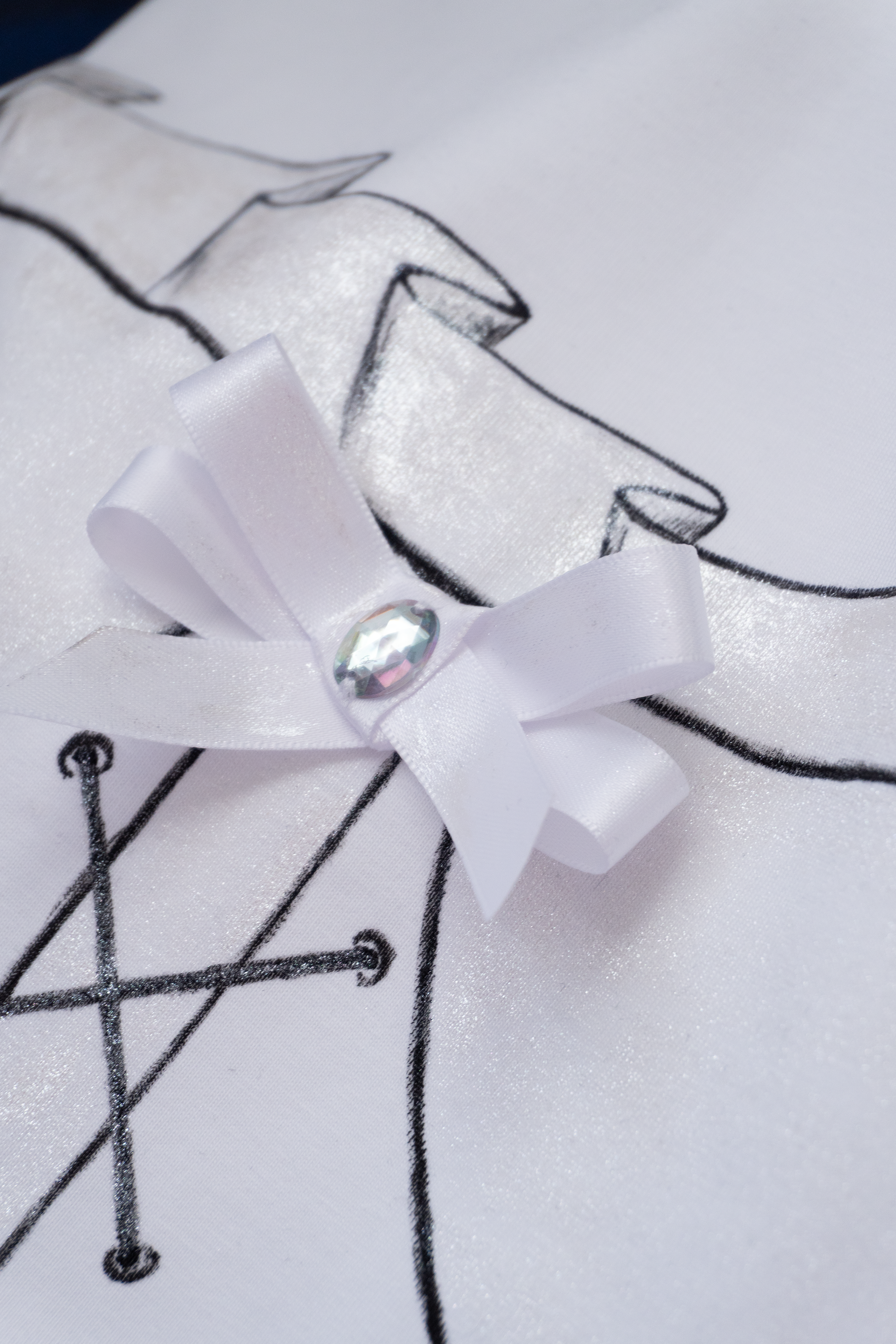 Close up detail of a white bow with a rhinestone in the centre of a hand painted corset on a white cotton tshirt and shimmery painted details