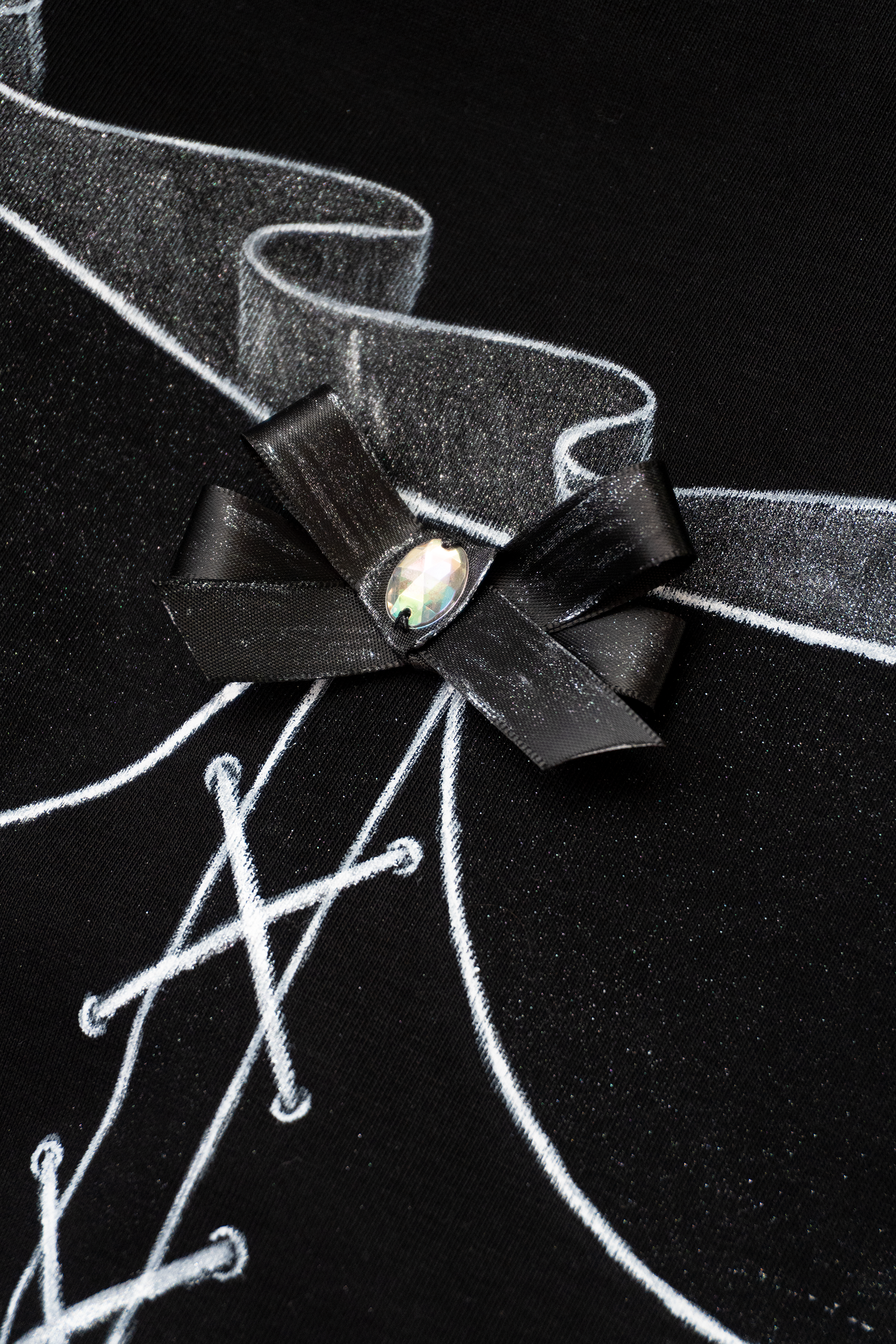 Close up detail of a black bow with a rhinestone in the centre of a hand painted corset on a black cotton tshirt and shimmery painted details